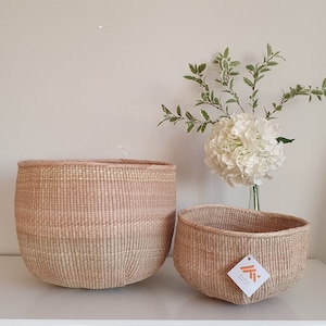 Puede incluir: Dos cestas tejidas, una grande y otra pequeña, hechas de materiales naturales. La cesta más grande es de color marrón claro y tiene una forma redondeada. La cesta más pequeña es de un color similar y tiene una forma ligeramente más redondeada. Ambas cestas están sobre una superficie blanca.