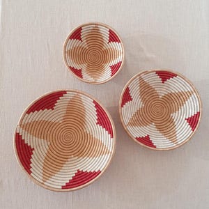 May include: Three woven baskets with a starburst design in shades of red, white, and beige. The baskets are arranged in a size order from largest to smallest.