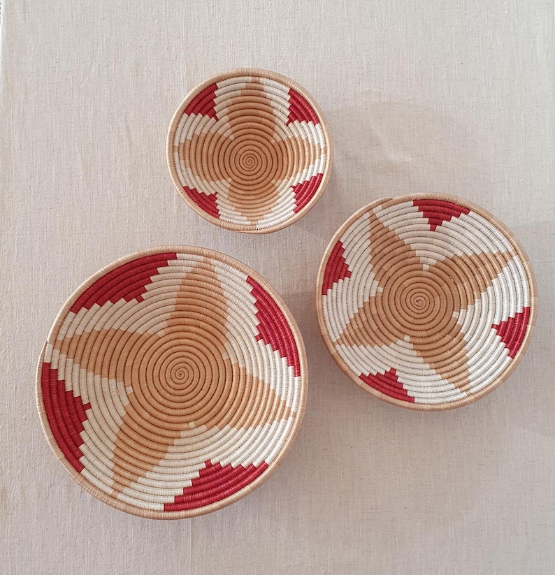 May include: Three woven baskets with a starburst design in shades of red, white, and beige. The baskets are arranged in a size order from largest to smallest.