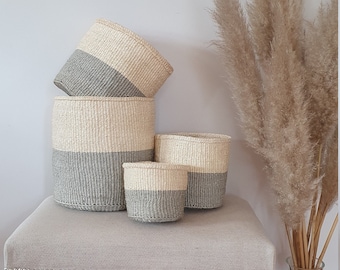 Puede incluir: Un conjunto de cuatro cestas tejidas de diferentes tamaños, cada una con una mitad superior beige natural y una mitad inferior gris. Las cestas están dispuestas sobre un banco tapizado gris claro, con hierba ornamental seca alta en el fondo.