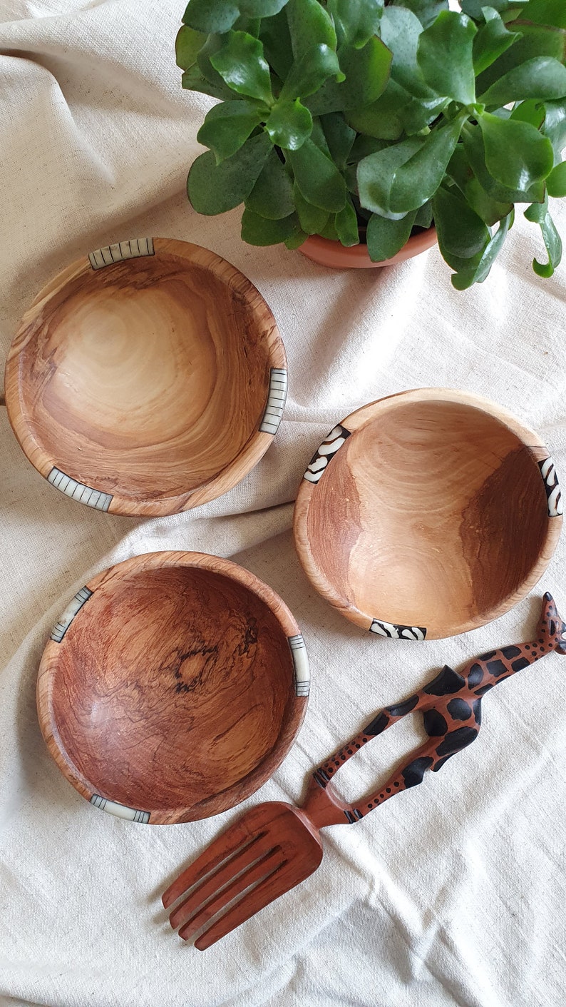Puede incluir: Tres cuencos de madera con incrustaciones decorativas, uno con un patr&oacute;n de cebra, otro con un patr&oacute;n de rayas y otro con un patr&oacute;n marr&oacute;n y blanco. Tambi&eacute;n se muestra un utensilio de servir de madera con forma de jirafa.
