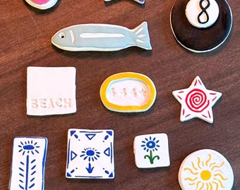 Könnte beinhalten: Eine Sammlung farbenfroher dekorativer Keramikmagnete. Designs umfassen einen Fisch, Sterne, eine Achtkugel und Quadrate mit floralen und geometrischen Mustern. Das Wort "BEACH" ist ebenfalls abgebildet.