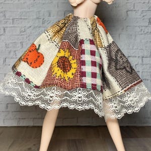 May include: A doll skirt featuring a patchwork design with autumn-themed patterns. The skirt includes a sunflower, pumpkin, and checkered fabric. The hem is trimmed with white lace. The skirt is designed for a doll.