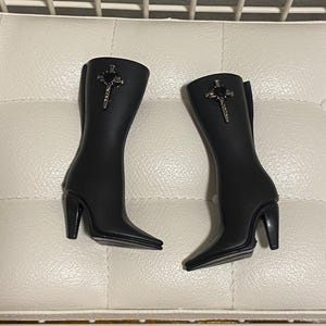 Puede incluir: Un par de botas negras de punta fina con tacón alto. Cada bota presenta un diseño de cruz y corazón plateado en la parte superior de la caña. Las botas son de material liso y mate.