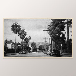 May include: A black and white photograph of a street lined with palm trees and cars. The Hollywood sign is visible in the distance.