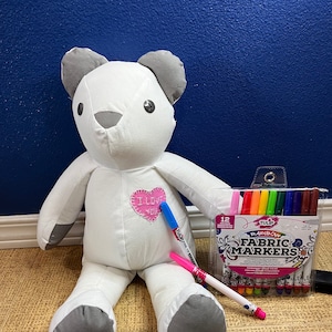 Può includere: Un orsacchiotto di peluche bianco con orecchie e zampe grigie è seduto su un pavimento di piastrelle marroni. L'orso ha un cuore rosa cucito sul petto con le parole "Ti amo" ricamate su di esso. Un pennarello per tessuti rosa e blu è accanto all'orso. Un pacchetto di 12 pennarelli per tessuti arcobaleno Tulip è sullo sfondo.