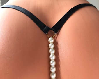 May include: A black elastic thong strap connected to a gold-toned metal ring, which serves as an anchor for a vertical strand of ten smooth, white, spherical faux pearls that extend downwards in a straight line against the skin.