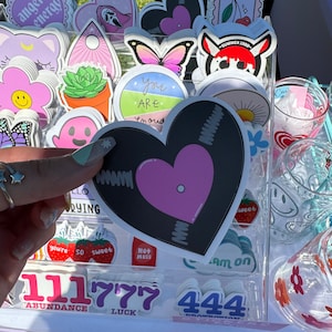 May include: Assortment of colorful stickers featuring various designs, including a heart-shaped record with a pink center, a butterfly, a succulent plant in a pot, a mushroom, and a sun. The stickers are arranged in a clear acrylic display case. The text "You Are You" is visible on one of the stickers.