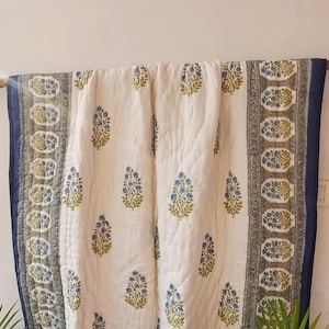 May include: A white cotton quilt with a blue and green floral pattern. The quilt is hanging on a white wall and has a blue border.
