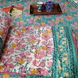 Pu&ograve; includere: Un piumino di cotone bianco con stampa floreale rosa e gialla. Il piumino &egrave; piegato a met&agrave; e ha un bordo turchese. Il piumino &egrave; su un letto con un copriletto a stampa floreale turchese.