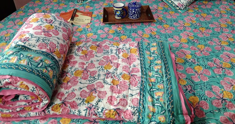 Pu&ograve; includere: Un piumino di cotone bianco con stampa floreale rosa e gialla. Il piumino &egrave; piegato a met&agrave; e ha un bordo turchese. Il piumino &egrave; su un letto con un copriletto a stampa floreale turchese.