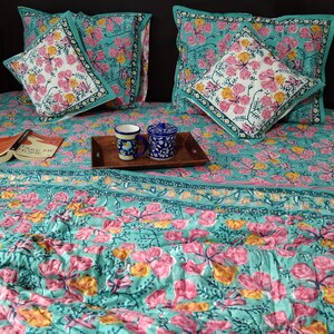 Pu&ograve; includere: Un copriletto con motivo floreale blu turchese e rosa con due federe abbinate e un cuscino pi&ugrave; piccolo con lo stesso motivo. Il copriletto ha un bordo a righe blu turchese e rosa. Un libro intitolato "Take Me Home" &egrave; appoggiato sul copriletto. Due tazze in ceramica blu e bianca sono su un vassoio di legno.