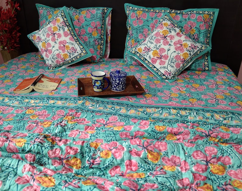 Pu&ograve; includere: Un copriletto con motivo floreale blu turchese e rosa con due federe abbinate e un cuscino pi&ugrave; piccolo con lo stesso motivo. Il copriletto ha un bordo a righe blu turchese e rosa. Un libro intitolato "Take Me Home" &egrave; appoggiato sul copriletto. Due tazze in ceramica blu e bianca sono su un vassoio di legno.