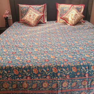 May include: A teal blue bedspread with a floral pattern featuring small red and orange flowers. The bedspread has a red and white border. Two red and white patterned pillows are on the bed.