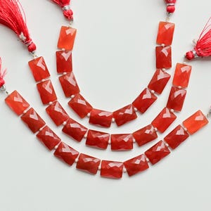 Puede incluir: Tres hilos de cuentas de piedras preciosas rojas con borlas rojas a juego. Las cuentas rectangulares tienen superficies facetadas y las borlas están hechas de hilos finos. Las cuentas están dispuestas en un patrón curvo sobre un fondo blanco.