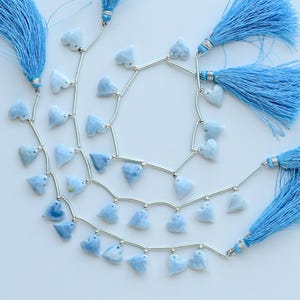May include: Three strands of light blue heart-shaped beads interspersed with thin silver-toned metal tubes. Each strand terminates with a long, light blue fabric tassel featuring a silver-toned metal cap at the top of the tassel.