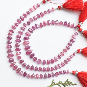 Puede incluir: Tres hebras de cuentas de piedras preciosas rosadas y moradas con superficies facetadas. Las cuentas están enhebradas en alambre plateado y tienen borlas rojas en cada extremo.