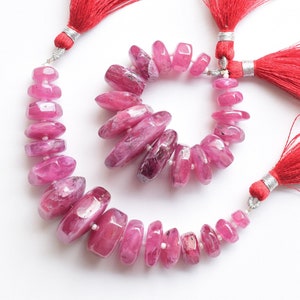 Puede incluir: Una hebra de cuentas de rubí rosa con forma alargada facetada. Las cuentas están enhebradas en un cordón plateado y tienen borlas rojas en cada extremo.