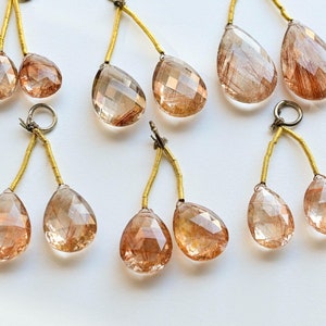 Könnte beinhalten: Acht tropfenförmige, facettierte, klare Quarzkristall-Anhänger mit goldfarbenen Drahtschlaufen. Die Kristalle haben eine rotbraune Farbe und enthalten Einschlüsse von goldenem Rutil.