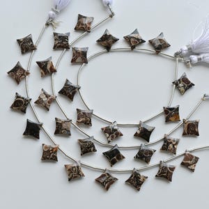 Pode incluir: Fios de contas em forma de estrela em tons de castanho, preto e creme, enfiadas em fio prateado. Tassels brancos adornam as extremidades. As contas apresentam um design único.