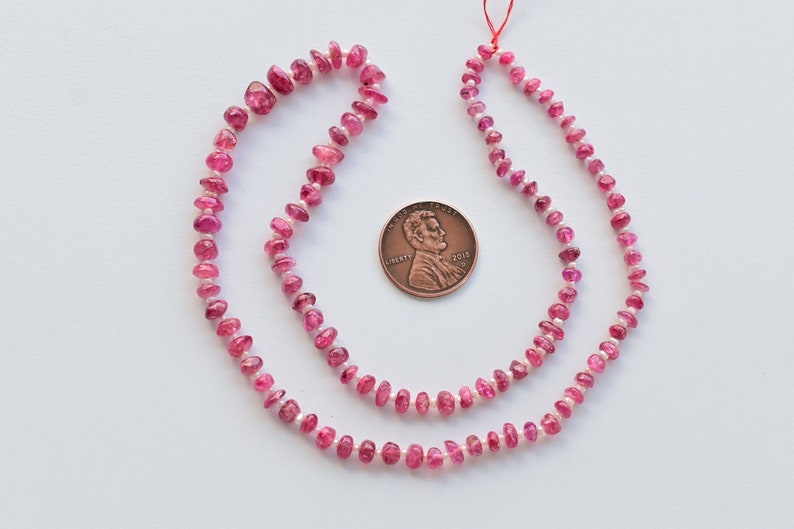 May include: A strand of pink tourmaline beads, approximately 16 inches long, with a penny for scale. The beads are faceted and have a slightly irregular shape.