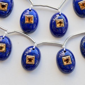 Könnte beinhalten: Eine Kette aus Lapislazuli-Perlen mit goldfarbenen Akzenten. Jede Perle ist oval geformt und hat einen quadratischen, goldfarbenen Stein in der Mitte.