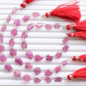 Puede incluir: Una hebra de cuentas de piedras preciosas rosadas con bordes facetados, enhebradas en alambre de plata con anillos de salto de plata. Las cuentas están dispuestas en un patrón circular, con borlas rojas unidas a los extremos de la hebra.