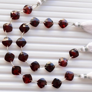 May include: A strand of dark red gemstone beads, each bead is a square shape, strung on silver wire with silver connectors. The strand is finished with white tassels on each end.