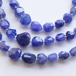 May include: Three strands of faceted, blue-violet tanzanite gemstone beads. The beads are irregularly shaped and strung on a white cord. The gemstones have a polished, reflective surface, and the background is a soft white.