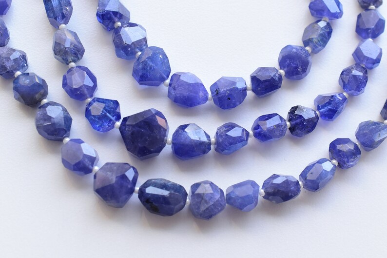 May include: Three strands of faceted, blue-violet tanzanite gemstone beads. The beads are irregularly shaped and strung on a white cord. The gemstones have a polished, reflective surface, and the background is a soft white.