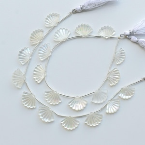 Könnte beinhalten: Zwei Stränge irisierender, fächerförmiger Perlen mit perlmuttartigem Glanz. Jede Perle ist auf einen silberfarbenen Draht gefädelt, und die Stränge sind mit weißen Quasten verziert. Die Perlen liegen auf einer weißen Oberfläche.