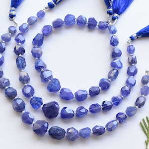 May include: Three strands of faceted, blue-violet tanzanite gemstone beads. Each strand is strung with small silver beads and finished with a blue tassel. The beads have an irregular, organic shape and are displayed on a white surface.