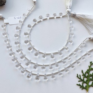 Puede incluir: Tres hebras de cuentas de cristal transparente en forma de lágrima enhebradas en alambre plateado. Las cuentas son facetadas y tienen una punta puntiaguda. Las hebras están dispuestas en círculo, con la hebra más grande en el exterior y la hebra más pequeña en el interior. El texto "ST-2" es visible en una etiqueta blanca.