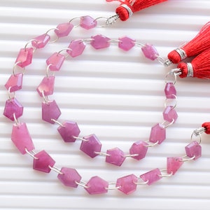 Puede incluir: Una hebra de cuentas de piedras preciosas rosas con conectores plateados. Las cuentas son facetadas y tienen forma hexagonal. La hebra está enhebrada en una cadena plateada y tiene dos borlas rojas unidas a los extremos.