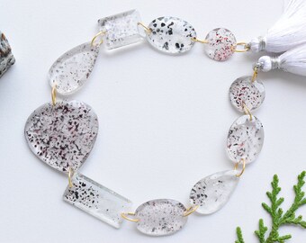 May include: A necklace featuring clear, irregularly shaped beads with black and red inclusions, strung together with gold-colored wire. The necklace is accented with white tassels. The beads vary in shape, including oval, teardrop, and rectangular forms.