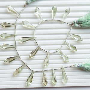 May include: A strand of light green gemstone beads with a faceted, teardrop shape. The beads are strung on a silver wire and have a tassel on each end.