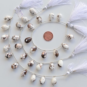 Puede incluir: Una hebra de cuentas de piedras preciosas blancas y marrones con detalles plateados y borlas blancas. Las cuentas son de forma ovalada y tienen un patrón de mármol. La hebra mide aproximadamente 45 cm de largo.