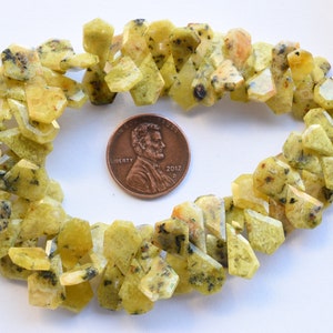 May include: A strand of yellow-green gemstone beads with a speckled pattern. The beads are faceted and have a rough, irregular shape. The beads are arranged in a circle around a UK penny for scale.