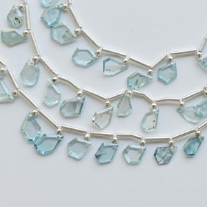 May include: Close-up of several strands of aquamarine gemstone beads. Each strand features irregular, faceted, light blue-green aquamarine stones strung on silver-colored wire. The beads are displayed against a white background.