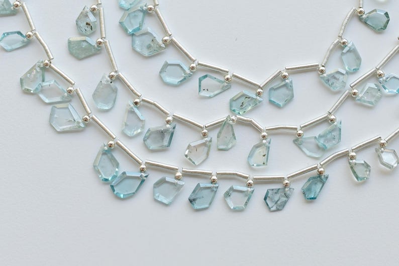 May include: Close-up of several strands of aquamarine gemstone beads. Each strand features irregular, faceted, light blue-green aquamarine stones strung on silver-colored wire. The beads are displayed against a white background.