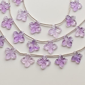 Könnte beinhalten: Eine Strähne aus Amethyst-Edelsteinperlen, jede in Form einer zarten Blume geschnitzt, auf Silberdraht gefädelt. Die Perlen sind hellviolett und haben eine durchscheinende Qualität.