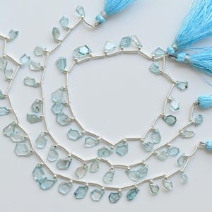 May include: Three strands of light blue gemstone beads with irregular shapes, strung on silver wire. The strands are accompanied by three light blue tassel accents, creating a layered and textured look. The beads are a pale, translucent blue.