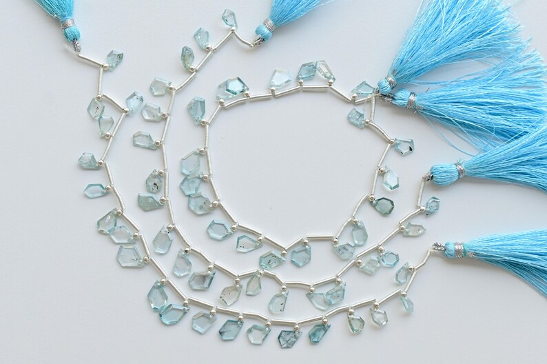 May include: Three strands of light blue gemstone beads with irregular shapes, strung on silver wire. The strands are accompanied by three light blue tassel accents, creating a layered and textured look. The beads are a pale, translucent blue.