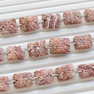 May include: A strand of square-shaped, pink and white patterned beads with carved floral designs. The beads are connected by silver jump rings.
