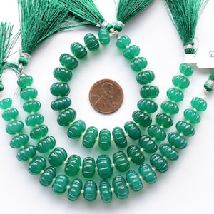 May include: A strand of green gemstone beads with a carved pumpkin shape. The beads are strung on a silver wire and have a green tassel at each end. The beads are approximately 10mm in diameter.