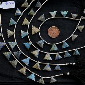 Puede incluir: Una hebra de cuentas de piedras preciosas triangulares de color azul y verde iridiscente en alambre plateado. Las cuentas están enhebradas en un patrón repetitivo y están separadas por pequeñas cuentas plateadas. La hebra se muestra sobre una superficie negra con una moneda de cobre y borlas negras.