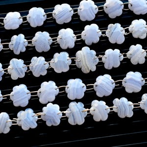 Pode incluir: Um fio de contas de ágata azul em forma de flor, conectadas por argolas de salto de prata. As contas têm um padrão listrado azul claro e branco.