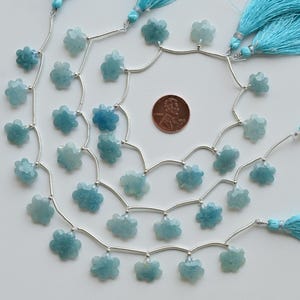 May include: Strands of light blue flower-shaped beads with silver wire and turquoise tassels. A penny is included for size reference. The beads are a pale blue color and have a faceted surface. The tassels are a bright turquoise color.