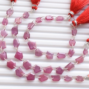 Puede incluir: Una hebra de cuentas de piedras preciosas rosadas con forma rectangular facetada. Las cuentas están enhebradas en alambre plateado y tienen un acabado pulido. La hebra está terminada con borlas rojas.