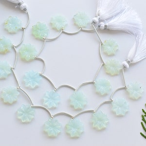 May include: A strand of light blue gemstone beads, each carved into a flower shape, strung on a silver chain with silver connectors. The chain has two white tassels attached to the ends.